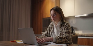 成熟的女商人在家工作，专注的女人坐在家里的厨房里用她的笔记本电脑和纸质文件工作。笔记本电脑应用程序计算成本，费用，估计预算的项目