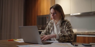 成熟的女商人在家工作，专注的女人坐在家里的厨房里用她的笔记本电脑和纸质文件工作。笔记本电脑应用程序计算成本，费用，估计预算的项目