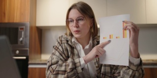 一位戴着眼镜的妇女坐在家里的厨房里，对着笔记本电脑的摄像头展示图形。家庭办公室远程工作。视频电话和会议与图形和表格。