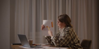 远程工作一位年轻女子戴着眼镜，坐在家里的厨房里进行在线报告，她正在展示笔记本电脑摄像头里的图形。视频电话和会议演示日程安排