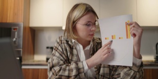 一位妇女坐在厨房里，对着笔记本电脑的摄像头展示图表。家庭办公室远程工作。视频电话和会议与图形和表格。解释图
