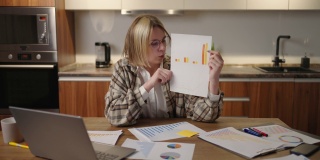 一位妇女坐在家里的厨房里，戴着眼镜，对着笔记本电脑的摄像头展示图表。家庭办公室远程工作。视频电话和会议与图形和表格。
