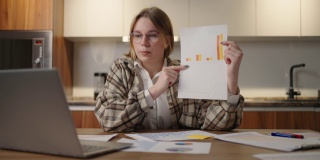 一位戴着眼镜的妇女坐在家里的厨房里，对着笔记本电脑的摄像头展示图表。家庭办公室远程工作。视频电话和会议与图形和表格。解释图