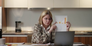 一位戴着眼镜的年轻女子坐在家里的厨房里，对着笔记本电脑的摄像头展示图表。家庭办公室远程工作。视频电话和会议与图形和表格。