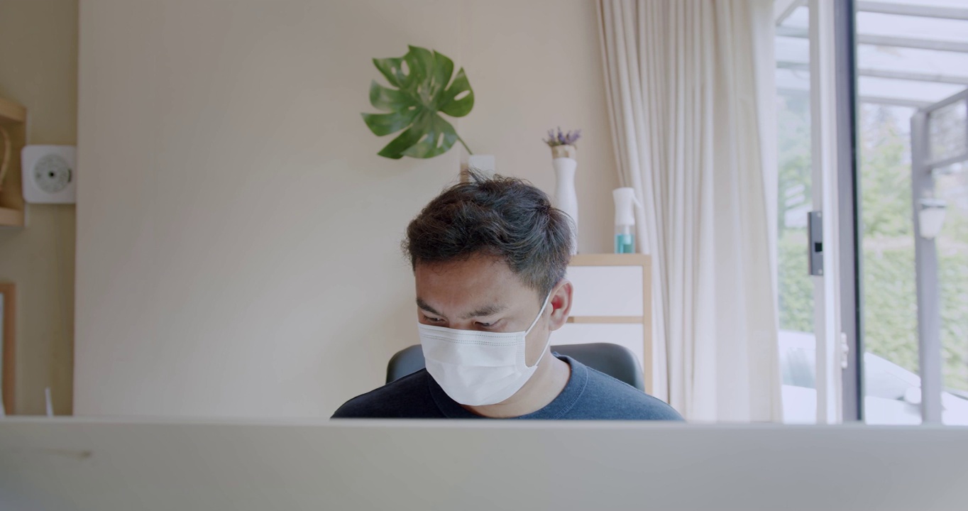 年轻的亚洲自由职业者男子戴着防护口罩在家里用电脑工作