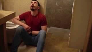 一个年轻的病人抱着自己的肚子坐在厕所旁边的地板上视频素材模板下载