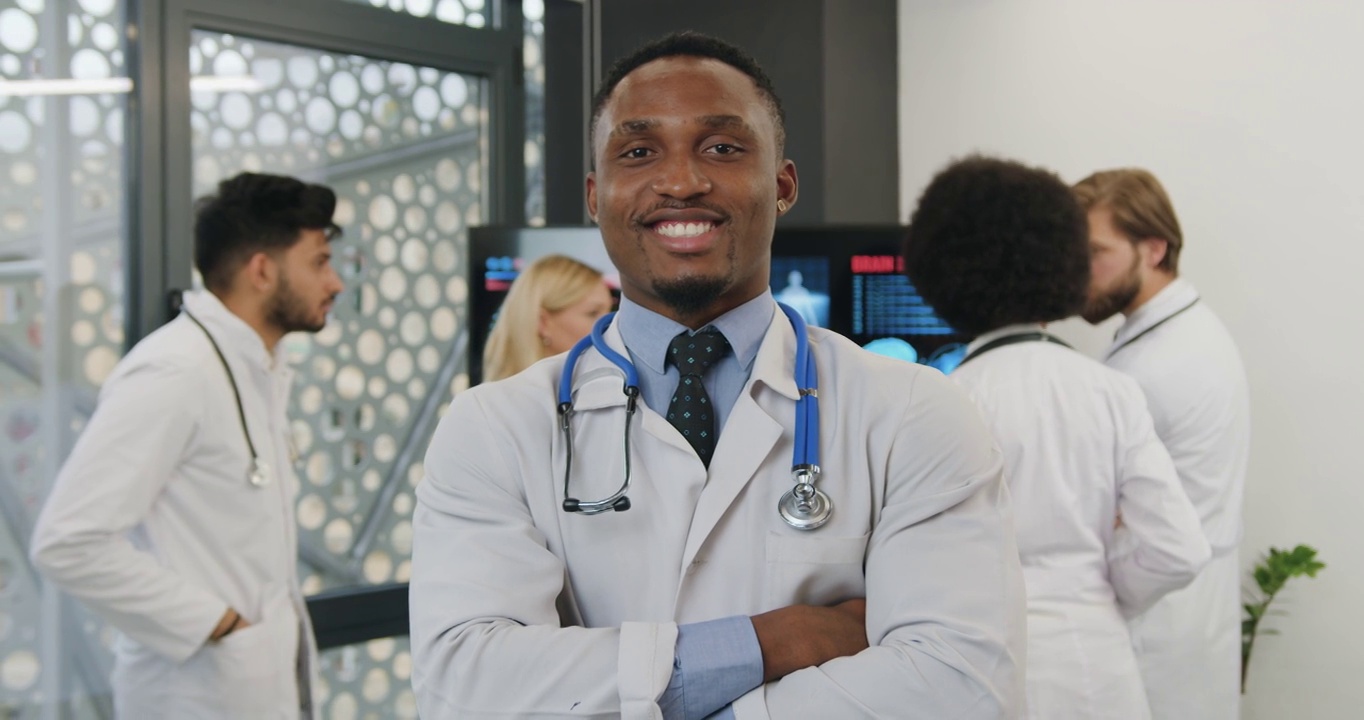 英俊、积极、友好、有经验的黑皮肤男性医生双臂交叉在镜头前摆姿势，而他的同事们则在数码显示器旁提供建议