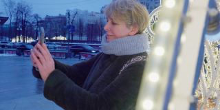 在圣诞节和新年期间，一个迷人的成熟女人，一名游客，在一个大城市用智能手机自拍和拍摄灯火通明的节日装饰。