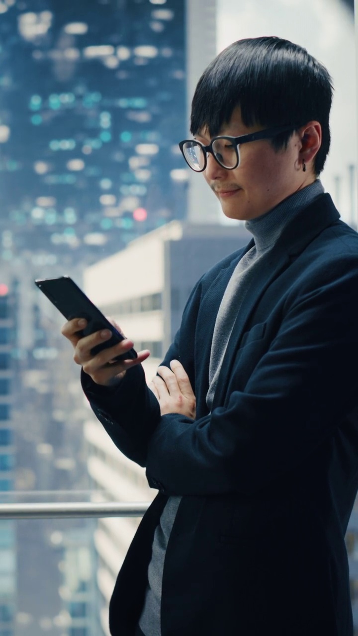 垂直屏幕:现代商务中心，时尚的日本商人乘坐玻璃电梯到办公室。帅哥在使用智能手机时微笑，写短信，在电梯里查看社交媒体。