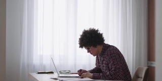卷发男学生英俊的年轻男孩戴着眼镜正在家里用笔记本电脑学习打字，在笔记本上写字。大学生使用笔记本电脑观看远程在线学习研讨会