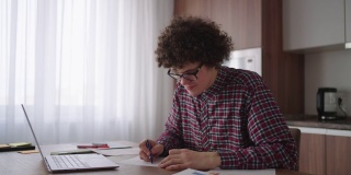 卷发男学生英俊的年轻男孩戴着眼镜正在家里用笔记本电脑学习打字，在笔记本上写字。大学生使用笔记本电脑观看远程在线学习研讨会