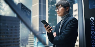 成功时尚的日本商人乘坐玻璃电梯到现代商务中心的办公室。帅哥在使用智能手机时微笑，写短信，在电梯里查看社交媒体。
