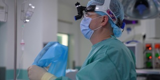 头上戴着特殊设备的男外科医生侧着身子站着。医生手里拿着创新的手术设备，聚精会神地直视前方。专业医生穿着特制制服，戴着帽子和口罩。