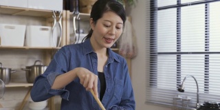 特写一个快乐的亚洲妇女品尝美味的食物从手指和扇手闻到香气，而在厨房做饭在家里。
