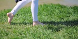 一个年轻女孩的脚赤脚走在绿色的草地在夏天一个阳光明媚的日子沿着湖。侧视图
