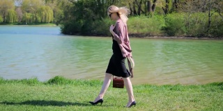 一位穿着办公服装的年轻女子，手里拿着手提包，戴着眼镜，心满意足地走在公园湖边的草地上。