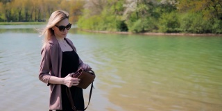 春天，一个年轻美丽的女人把一些东西放在她的手提包里，背景是城市公园里的一个湖