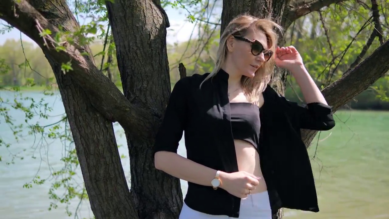 一位漂亮的年轻女子戴着太阳镜，穿着黑色的上衣和一件没有扣扣子的衬衫，站在树下，在湖边的大自然中拍照
