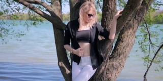 夏天，一位戴着墨镜、身穿黑色上衣和衬衫、身穿白色牛仔裤的金发女子站在公园湖边的一棵树旁