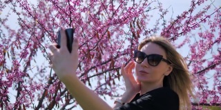 一位年轻美丽的女子在一棵盛开的粉红色樱花树的背景下自拍，欣赏自己