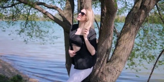 一个美丽的金发女人穿着短上衣，戴着太阳镜，在自然的湖边的一棵树旁摆出性感的姿势
