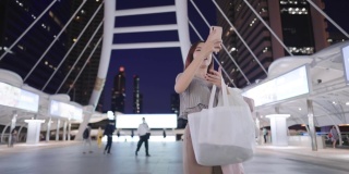 迷人的亚洲年轻成人视频通话，行走在现代化的市中心过桥夜间，愉快的女工作人员使用智能手机应用程序，在线互联网连接