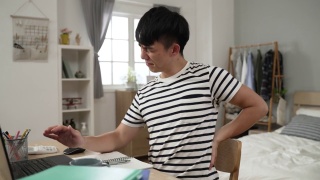 一名疲惫的亚洲大学生感到腰痛，一边在电脑上做作业，一边苦笑着给自己按摩视频素材模板下载