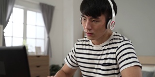 疲惫的亚洲男子成年学生脱下耳机，揉着疼痛的眼睛，白天在卧室的电脑上学习。