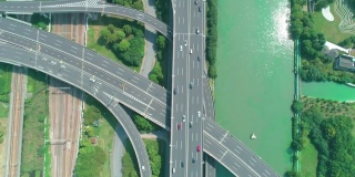 从空中俯瞰高速公路立交桥多层路口，快速移动的汽车被绿树环绕，在阳光明媚的一天，有一条河在一边