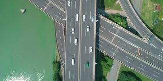 从空中俯瞰高速公路立交桥多层路口，快速移动的汽车被绿树环绕，在阳光明媚的一天，有一条河在一边。