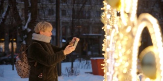 有吸引力的成熟女人，一个游客，在圣诞节期间在一个大城市享受节日装饰。她正在用智能手机自拍，转过身来拍照明照片。