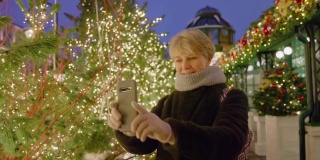 有魅力的成熟女子，一名游客，在圣诞节期间用智能手机在灯火通明的公园里拍照和自拍，享受大城市的节日装饰。
