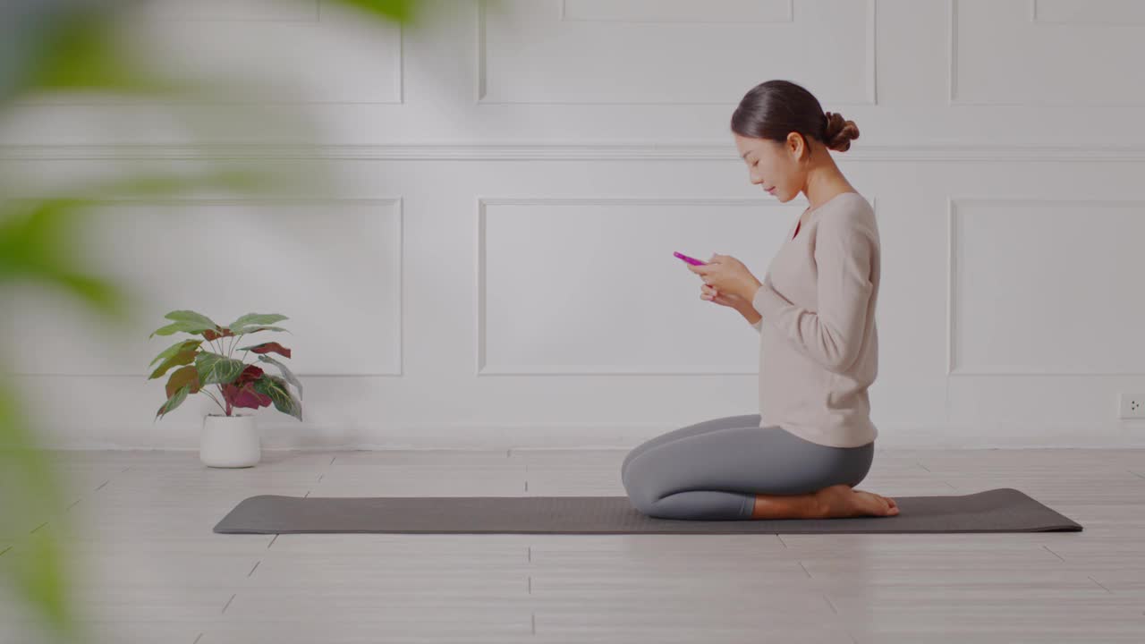 快乐的亚洲年轻女子坐在瑜伽垫放松与手机采取刹车后，与瑜伽练习在家里。瑜伽锻炼后休息的女人用手机发短信，面带微笑，心情愉悦