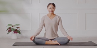平静的健康亚洲年轻女性坐在地毯上呼吸瑜伽莲花姿势，瑜伽冥想年轻健康女性放松和舒适的白色舒适的家，瑜伽锻炼为健康的概念
