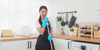 亚洲清洁服务女孩唱歌，而清洁厨房在家里。美丽的年轻快乐的女人管家清洁工跳舞和拖地工作，家务或家务在家里。