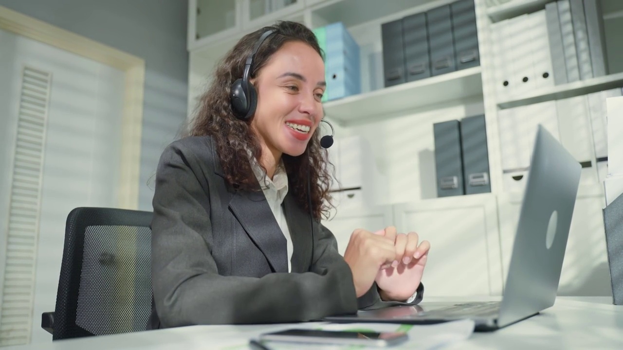 美丽的拉丁裔商务女性呼叫中心微笑，而工作在办公室。漂亮的年轻女员工穿着正装坐在工作场所的桌子上，用笔记本电脑交谈来支持客户。
