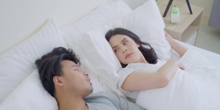 亚洲女性醒来后因丈夫打鼾而感到沮丧。美丽迷人的新婚夫妇，清晨在家里卧室的床上睡觉时，男人和女人有噪音。