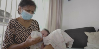 奶奶戴着口罩，用奶瓶喂孙子喝牛奶。在covid-19。正在吸奶的新生儿。