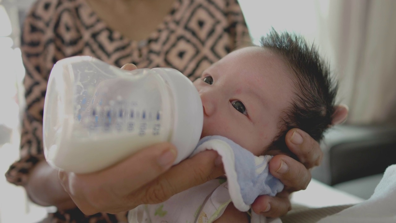 奶奶戴着口罩，用奶瓶喂孙子喝牛奶。在covid-19。正在吸奶的新生儿。
