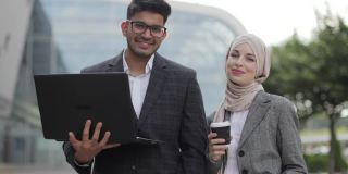 商务人士带着笔记本电脑和咖啡在户外。英俊的印度男子和穆斯林女士在商业中心外的相机前摆姿势，使用笔记本电脑和喝咖啡。