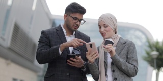 阿拉伯商务夫妇在茶歇时走在街上，谈笑风生。戴头巾的女人向她的同事展示她的智能手机屏幕，分享想法或新闻。