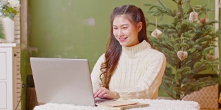 亚洲女商人工作呆在家里，圣诞树在背景庆祝工作成功。自由职业妇女在平安夜用电脑工作。假期工作概念