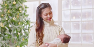 美丽的亚洲妇女抱着新生儿坐在白色舒适的家里的床上，背景是圣诞树。快乐的宝宝睡在妈妈的怀里，又安全又舒适。母婴概念