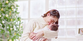 美丽的亚洲妇女抱着新生的婴儿，亲吻坐在白色舒适的家里的床上，背景是圣诞树。快乐的宝宝睡在妈妈的怀里，又安全又舒适