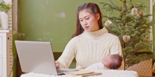 快乐的亚洲母亲妇女抱着她的婴儿新生儿和工作呆在家里。自由职业妈妈用电脑工作，在圣诞节前夕在家照顾她的新生儿