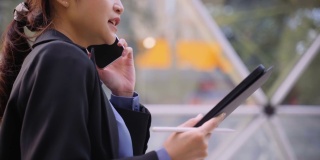 亚洲女商人使用笔记本电脑，在城市的办公室前用她的手机交谈