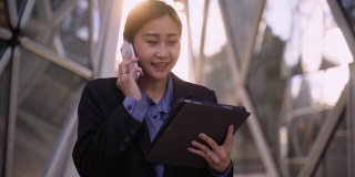 自信的亚洲女性企业家，在现代商业大厦附近一边走一边用数码平板电脑和电话聊天