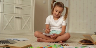 在家里上学的时候，一个快乐的小女孩坐在家里的地板上看着笔记本电脑屏幕写作。
