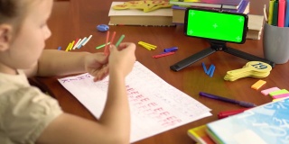 一个学龄前的小女孩展示了计数棒，看着一个绿色屏幕的智能手机。后视图。