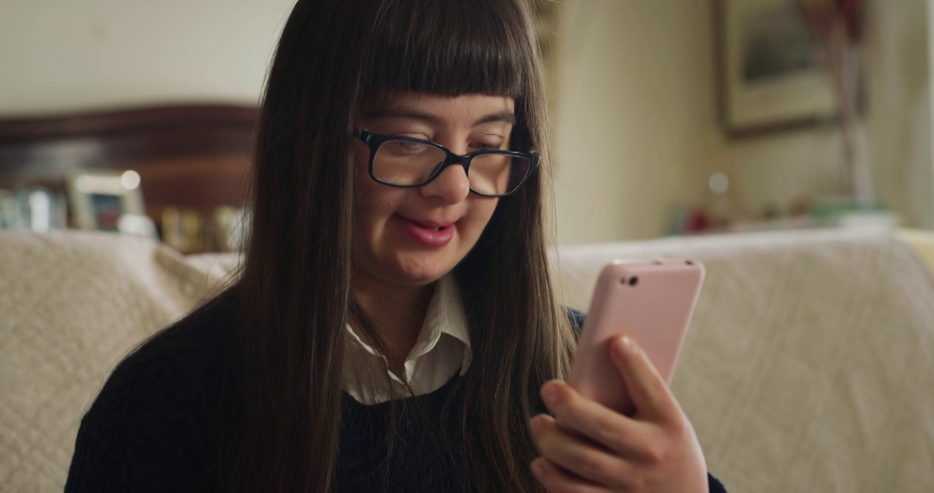 电影镜头快乐微笑的青少年女孩与唐氏综合症使用智能手机家庭娱乐在家里。概念技术，连接，残障，残障，在线，沟通，媒体